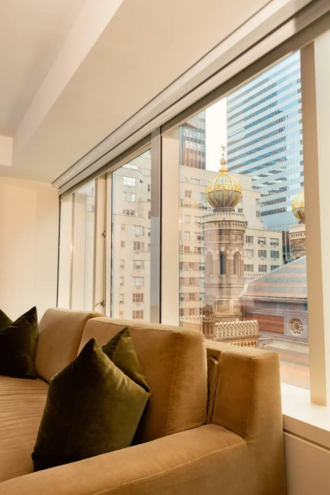 A beige sofa with green pillows sits by a large window offering a view of city skyscrapers and a historic gold-domed building.