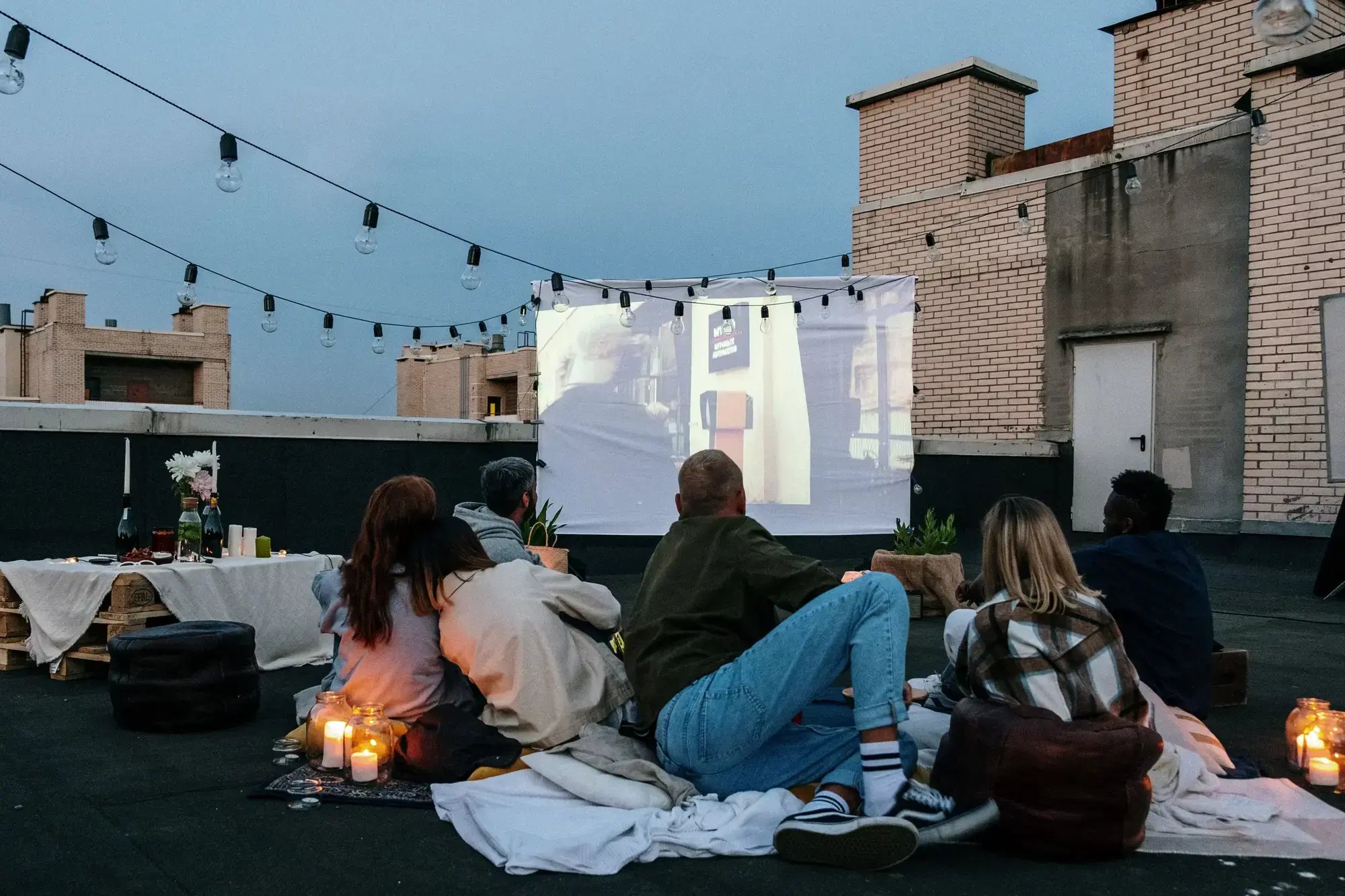 Friends enjoying a cozy rooftop movie night with candles and blankets under string lights