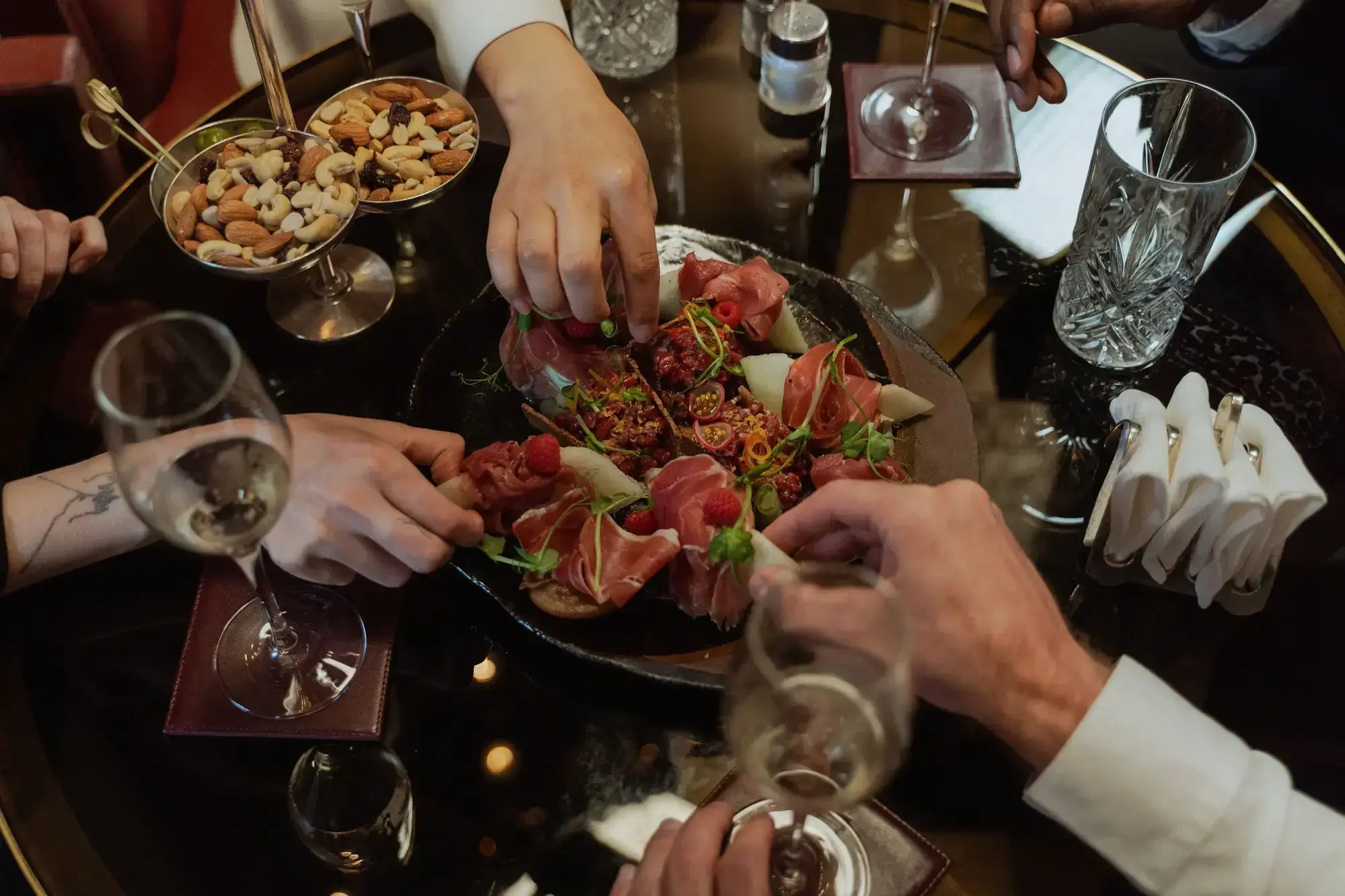 A sophisticated dining setting featuring prosciutto, wine, and mixed nuts on a glass table.