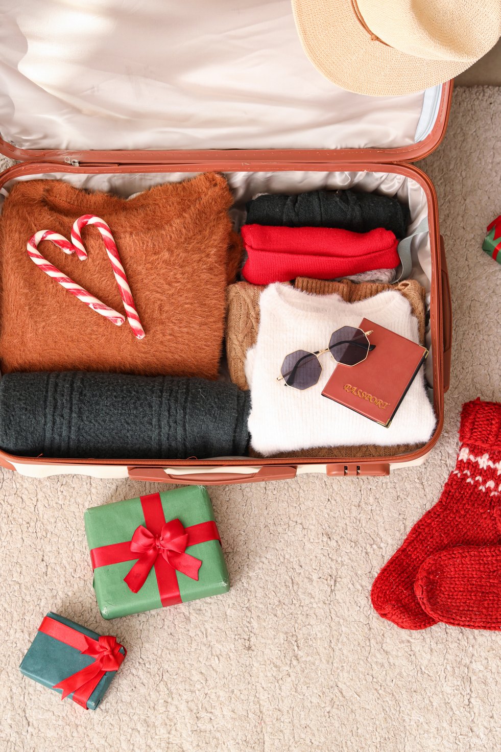 A suitcase packed with cozy winter clothes, candy canes arranged in a heart shape, a passport with sunglasses on top, and Christmas gifts and red knitted socks placed beside it.