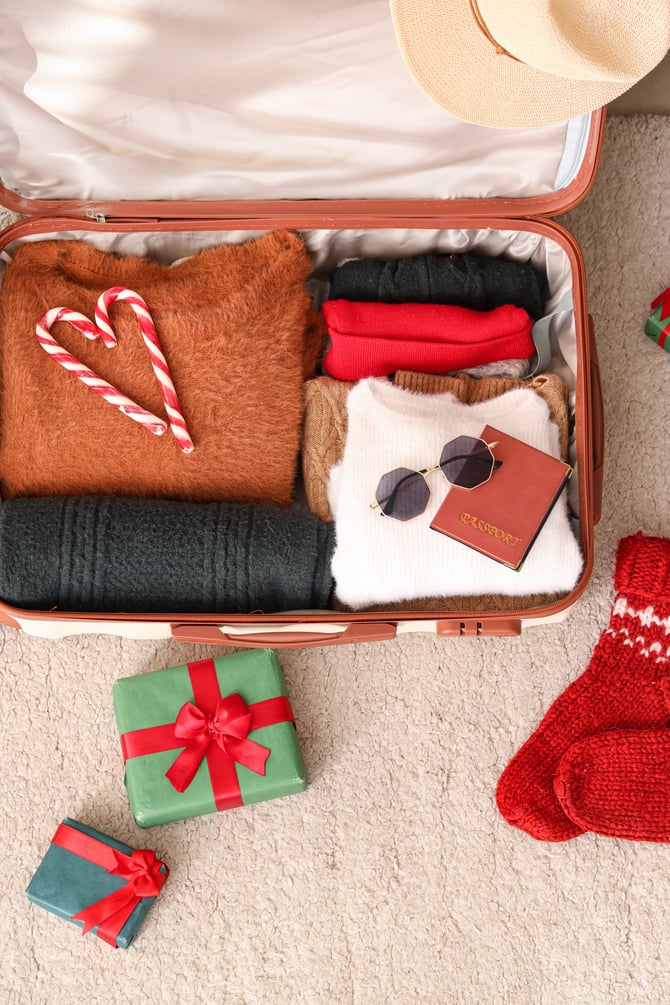 A suitcase packed with cozy winter clothes, candy canes arranged in a heart shape, a passport with sunglasses on top, and Christmas gifts and red knitted socks placed beside it.